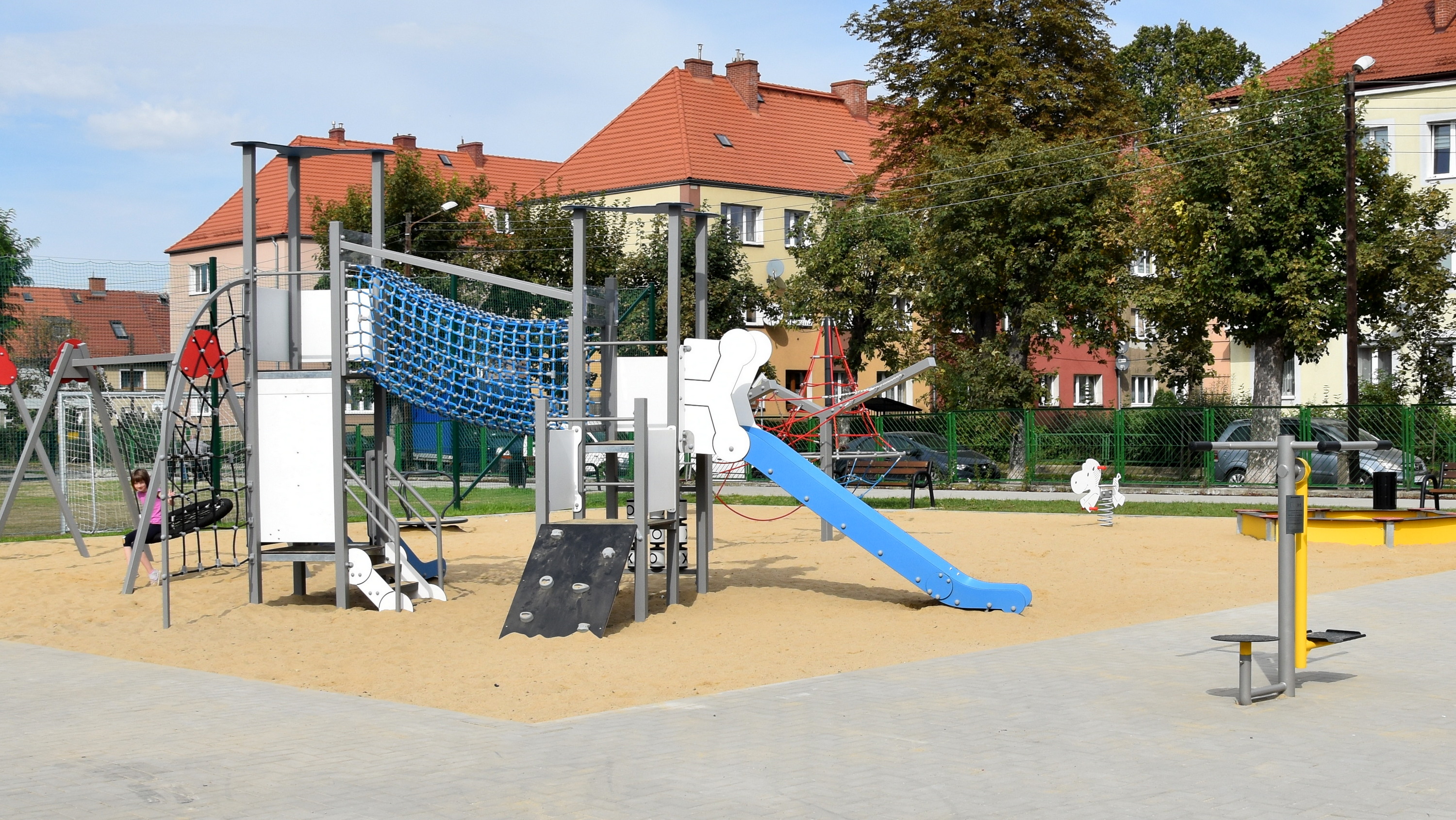 Nowy plac zabaw, piaszczyste podłoże, urządzenia do zabawy, w tle drzewa i budynki mieszkalne