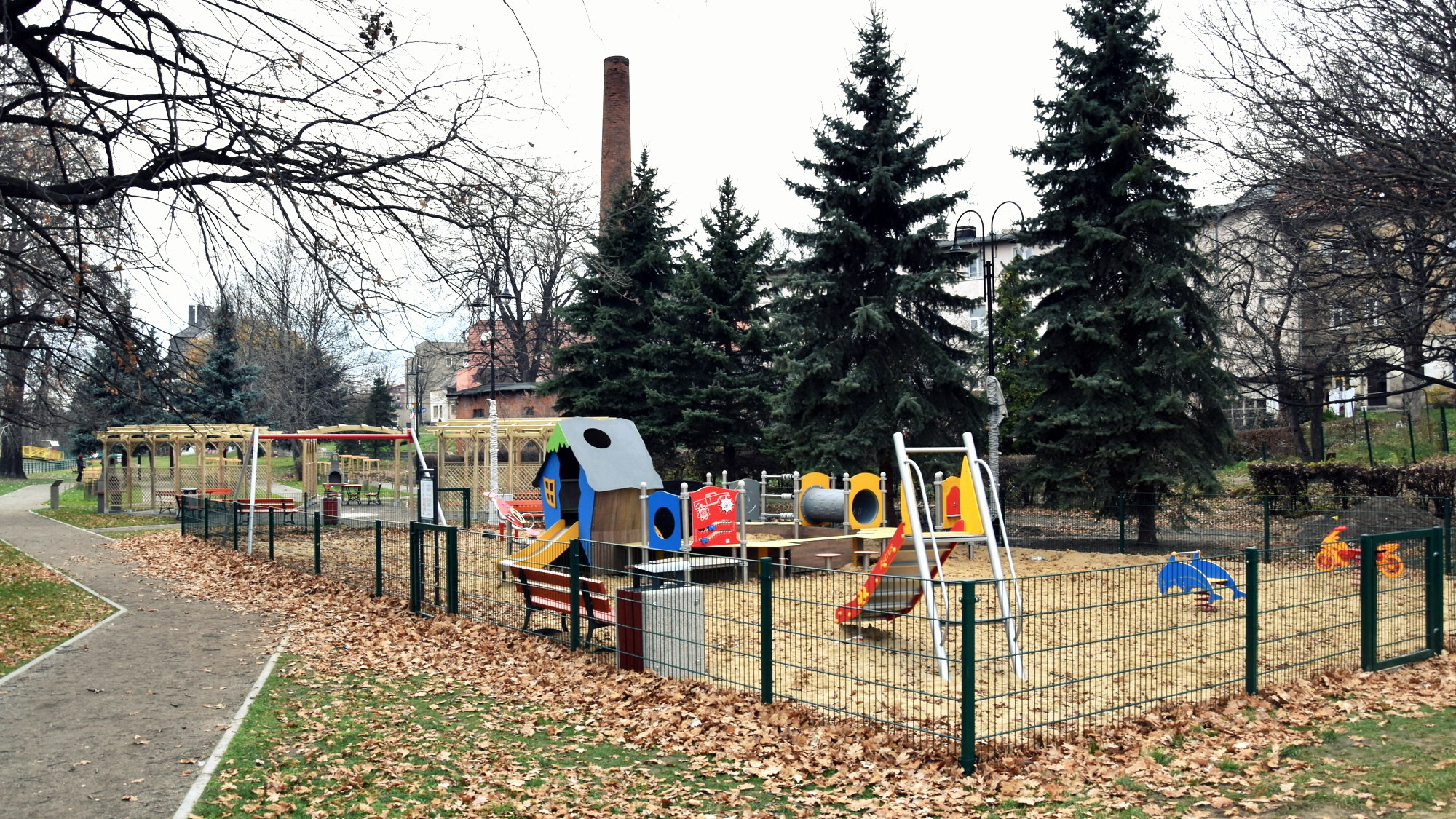 Plac zabaw z kolorowymi urządzeniami i piaskowym podłożem, w drugim planie wiaty grillowe