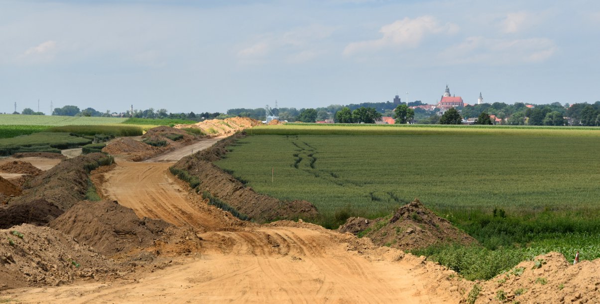 Droga w początku budowy, po bo bokach zielone pola, w dali panorama miasta