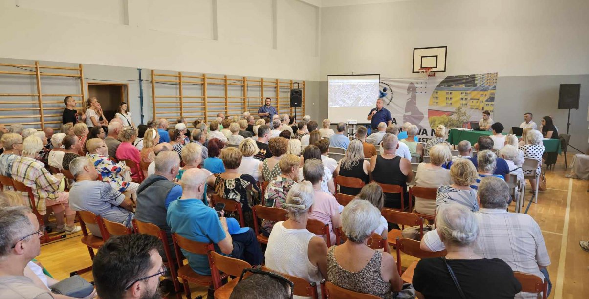 Sala gimnastyczna, ludzie siedzący na krzesłąch, w drugim planie burmistrz z mikrofonem