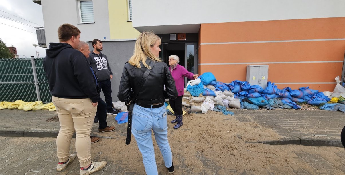 Grupa ludzi stojących przed budynkie, przy nim terta worków, ktłóra broniła przed powodzią