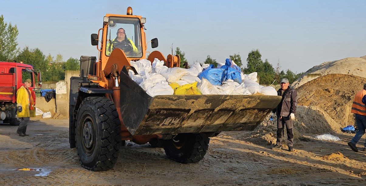 Ładowarka wiezie na "łyżce" worki z piaskiem, w tle cieżąrówka, ludzie i góry piachu