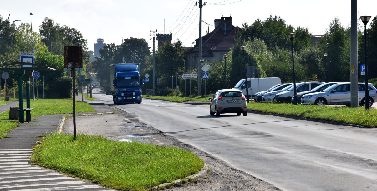 Droga wjazdowa do miasta, dwa jadące samochody, obok parking ze stojącymi autami