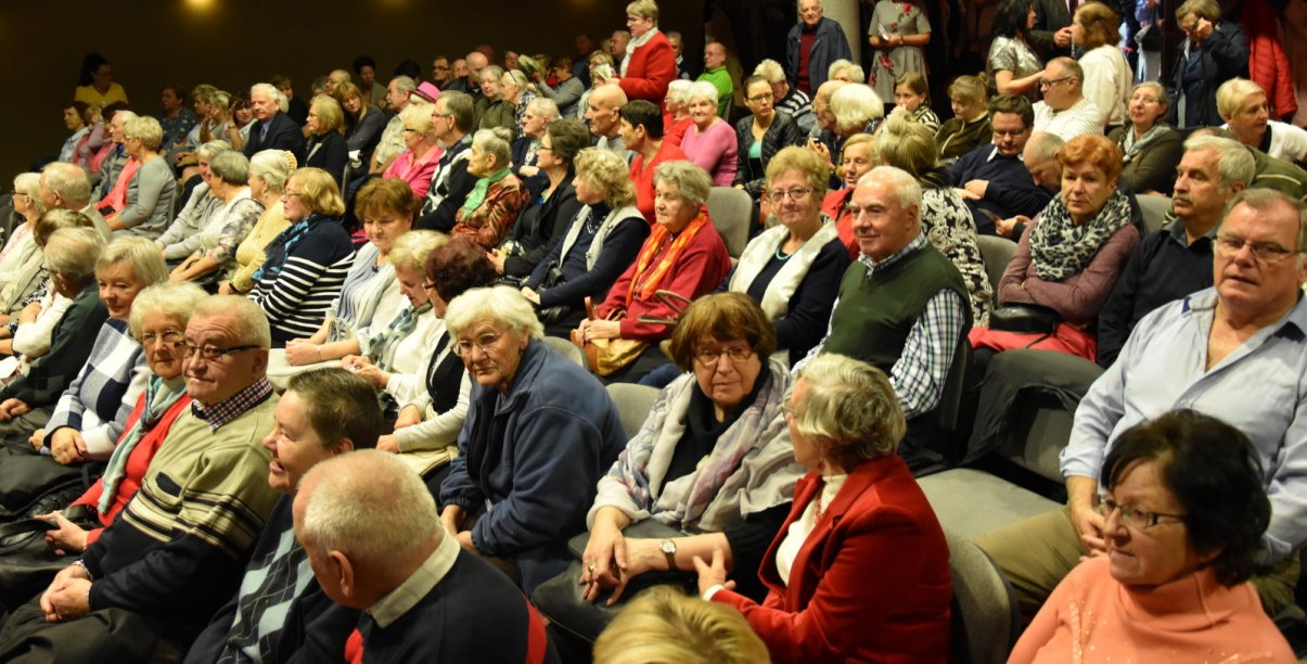 Kilkudziesięciu seniorów siedzących na krzesałach w sali ośrodka kultury