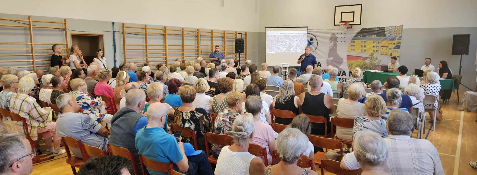 Sala gimnastyczna, ludzie siedzący na krzesłąch, w drugim planie burmistrz z mikrofonem