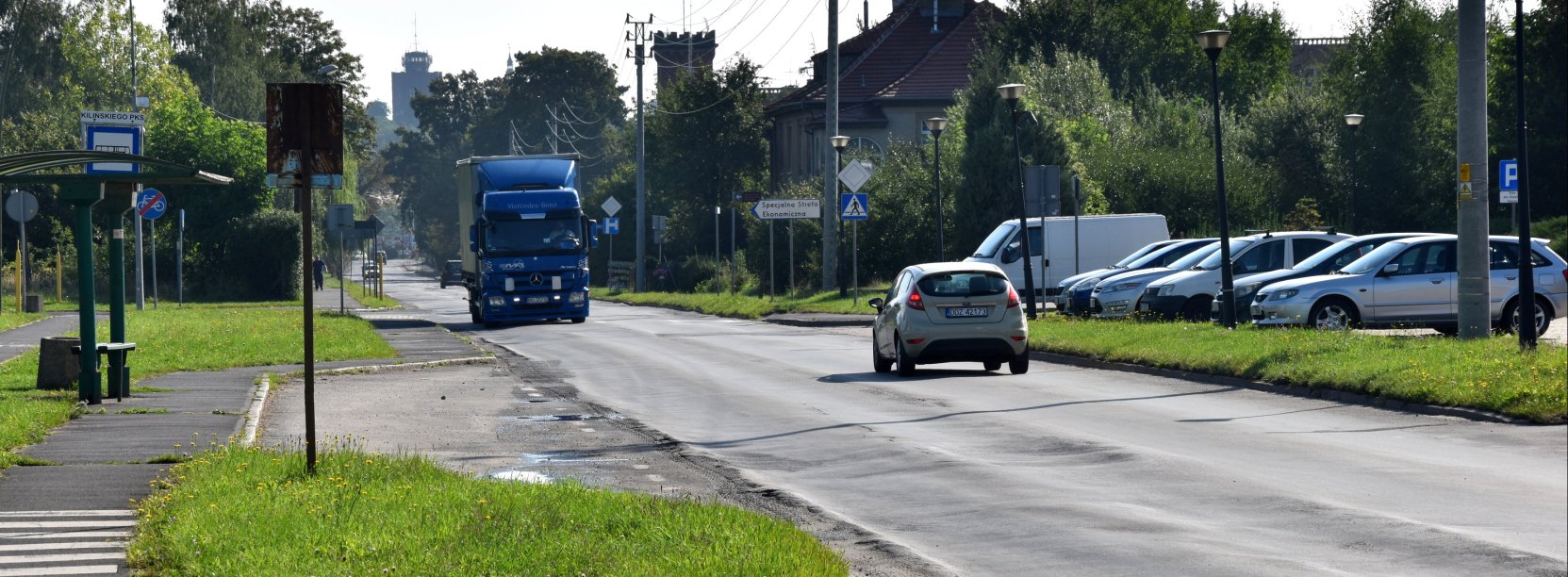 Droga wjazdowa do miasta, dwa jadące samochody, obok parking ze stojącymi autami