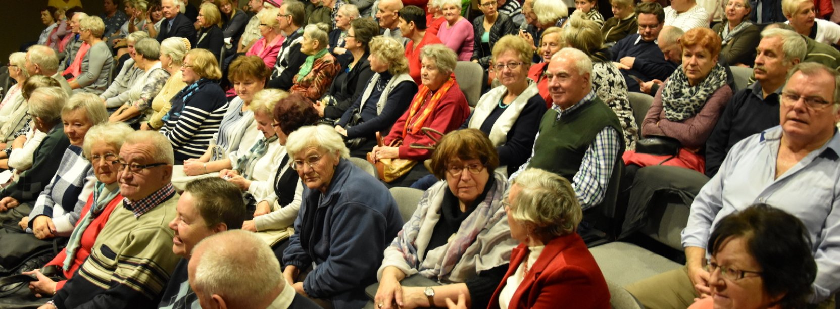 Kilkudziesięciu seniorów siedzących na krzesałach w sali ośrodka kultury