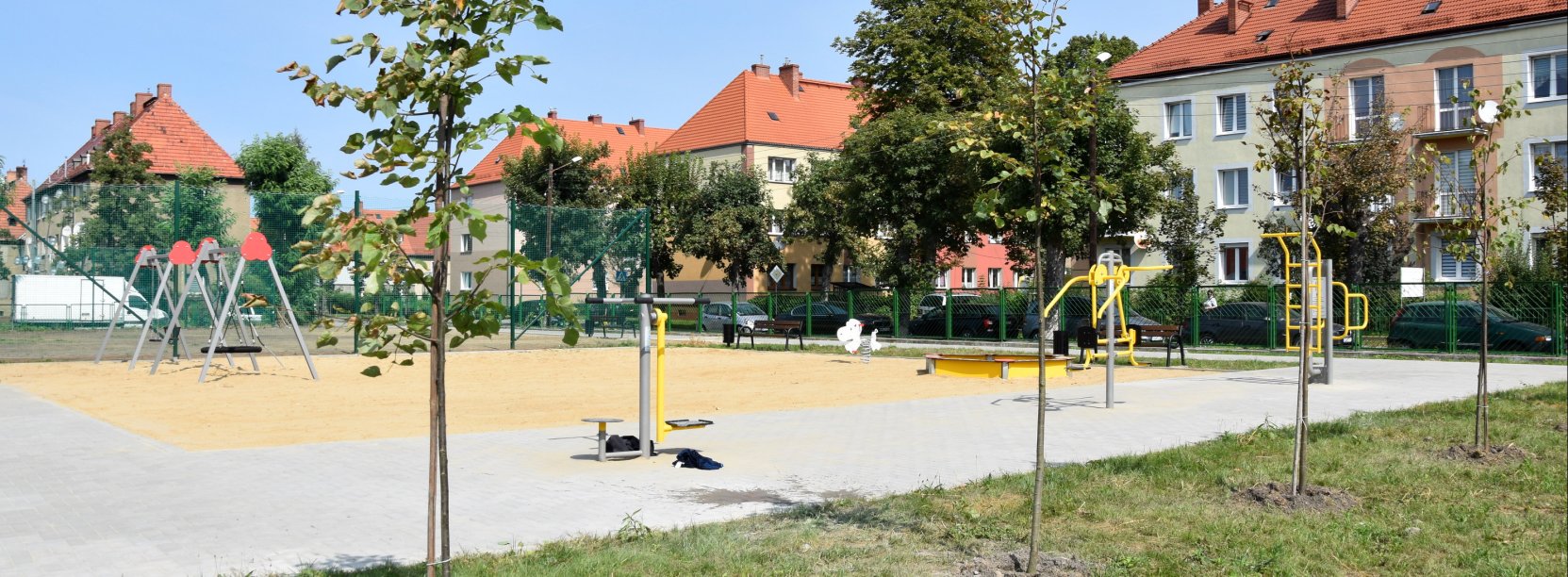 Plac zabaw z dużą piaskownicą, na pierwszym planie drzewka, w tle budynki mieszkalne