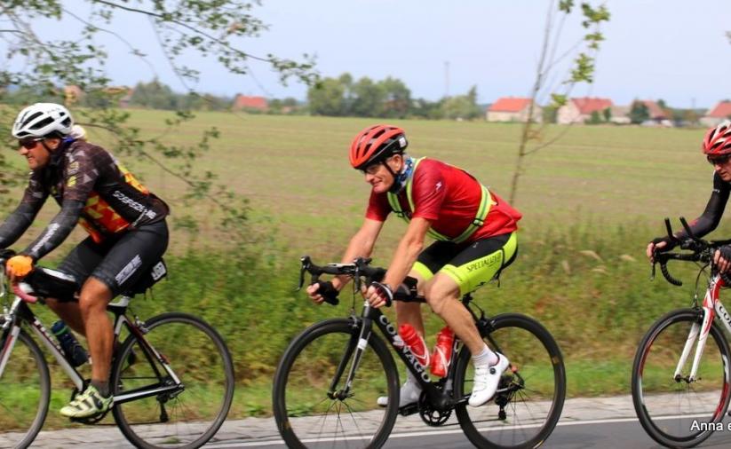 Trzech rowerzystów jadących po drodze, w drugim planie łąka i wiejskie zabudowy