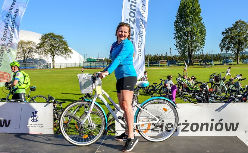 Uśmiechnięta kobieta na rowerze, za nią boisko na którym siedzi grupa rowerzystów