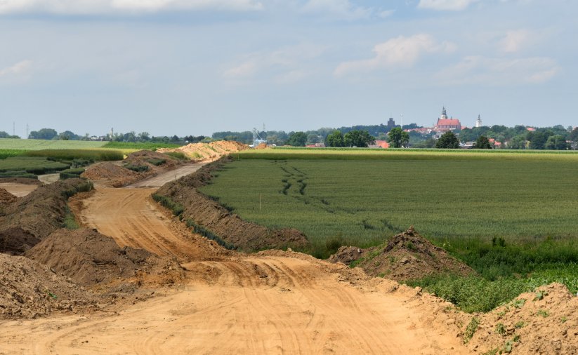 Droga w początku budowy, po bo bokach zielone pola, w dali panorama miasta