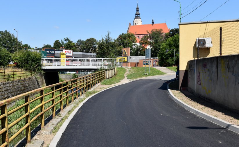 Droga z nowym asfaltem, po lewej kamienne koryto rzeki, po prawej mur, w drugim planie średniowieczny kościół