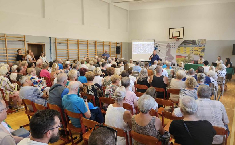 Sala gimnastyczna, ludzie siedzący na krzesłąch, w drugim planie burmistrz z mikrofonem