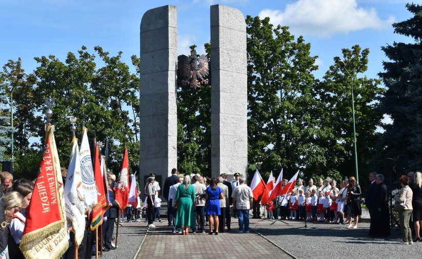 Grupa ludzi stojąca przed pomnikiem, obok poczet sztandarowy i młodzież w białych koszulach 