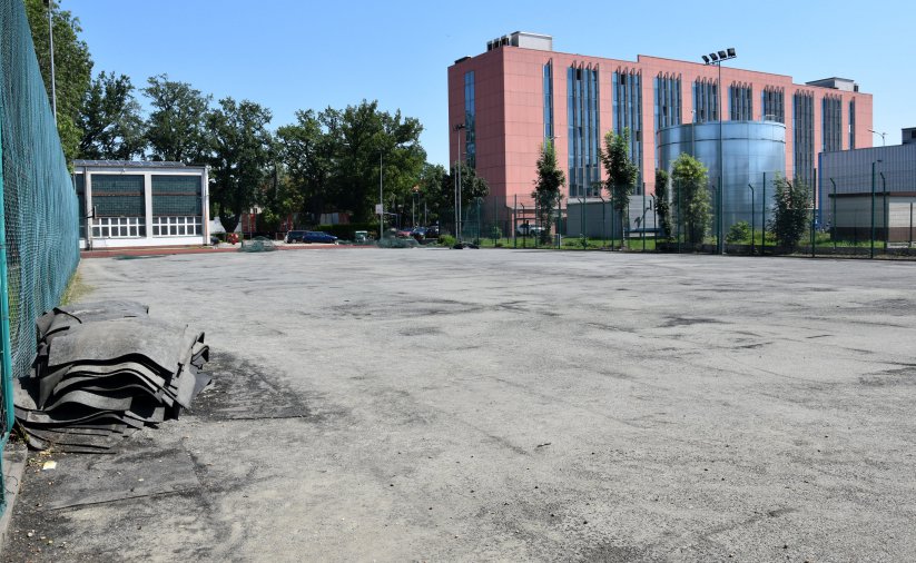 Boisko do piłki nożnej ze zdjętą sztuczną nawierzchnią, w drugim planie czerwony wielopiętrowy budynek