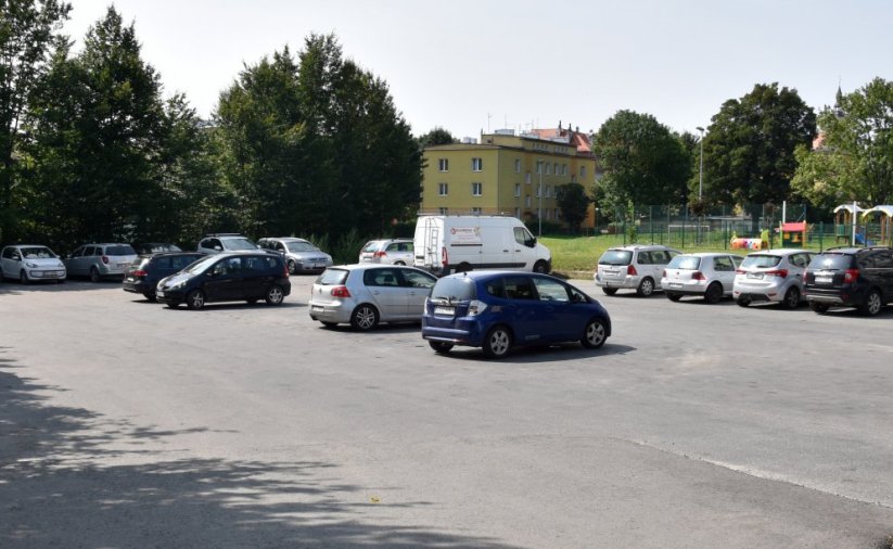 Asfaltowy parking z zaparkowanymi autami, po prawej stronie budynki