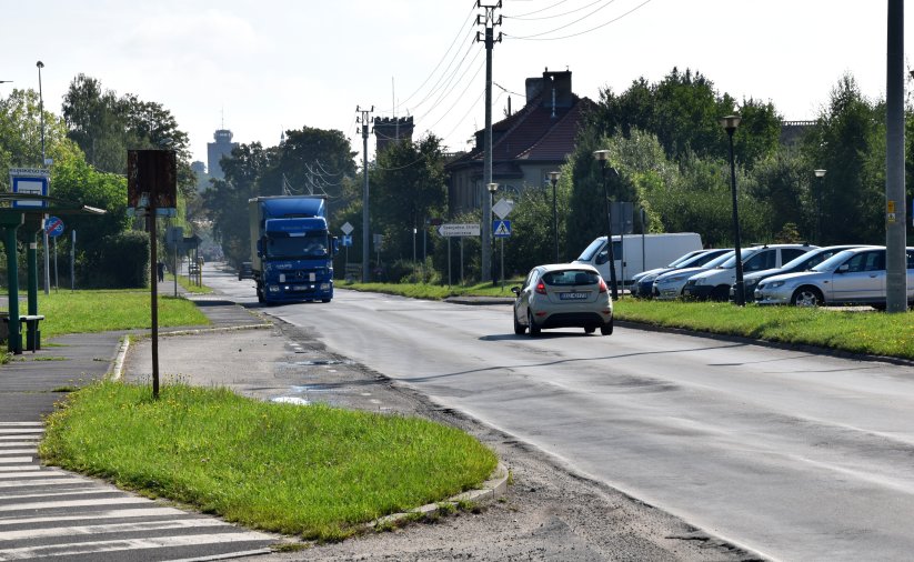 Droga wjazdowa do miasta, dwa jadące samochody, obok parking ze stojącymi autami