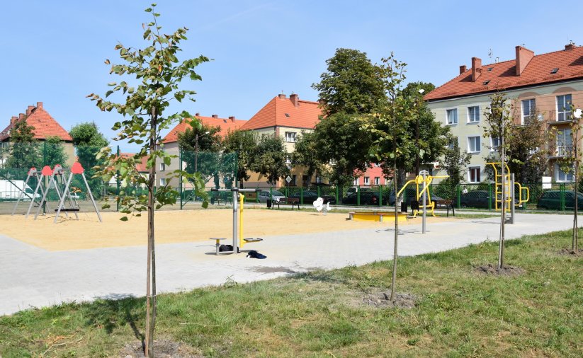 Plac zabaw z dużą piaskownicą, na pierwszym planie drzewka, w tle budynki mieszkalne