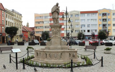 Rzeźba Jana Nepomucena na cokole, otoczaona kamiennymi barierkami, w tle budynki