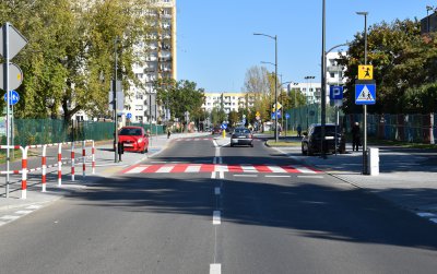 Droga osiedlowa z przejściami dla pieszych i miejscami postojowymi, w tle zieleń i bloki mieszkalne