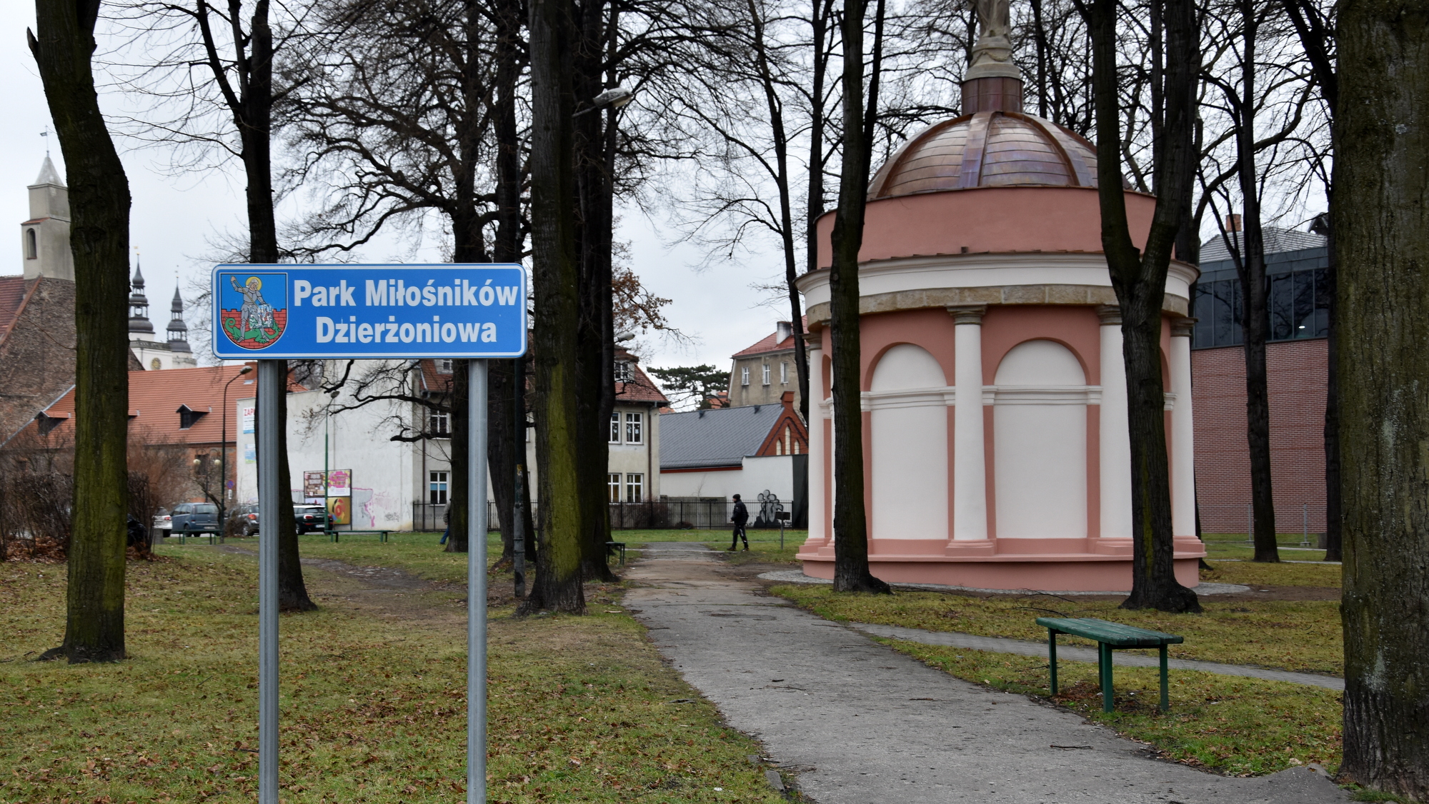 Park, po lewej tabica z nazwą Park Miłośników Dzirżowniowa, w drugim planie okrągła kaplica
