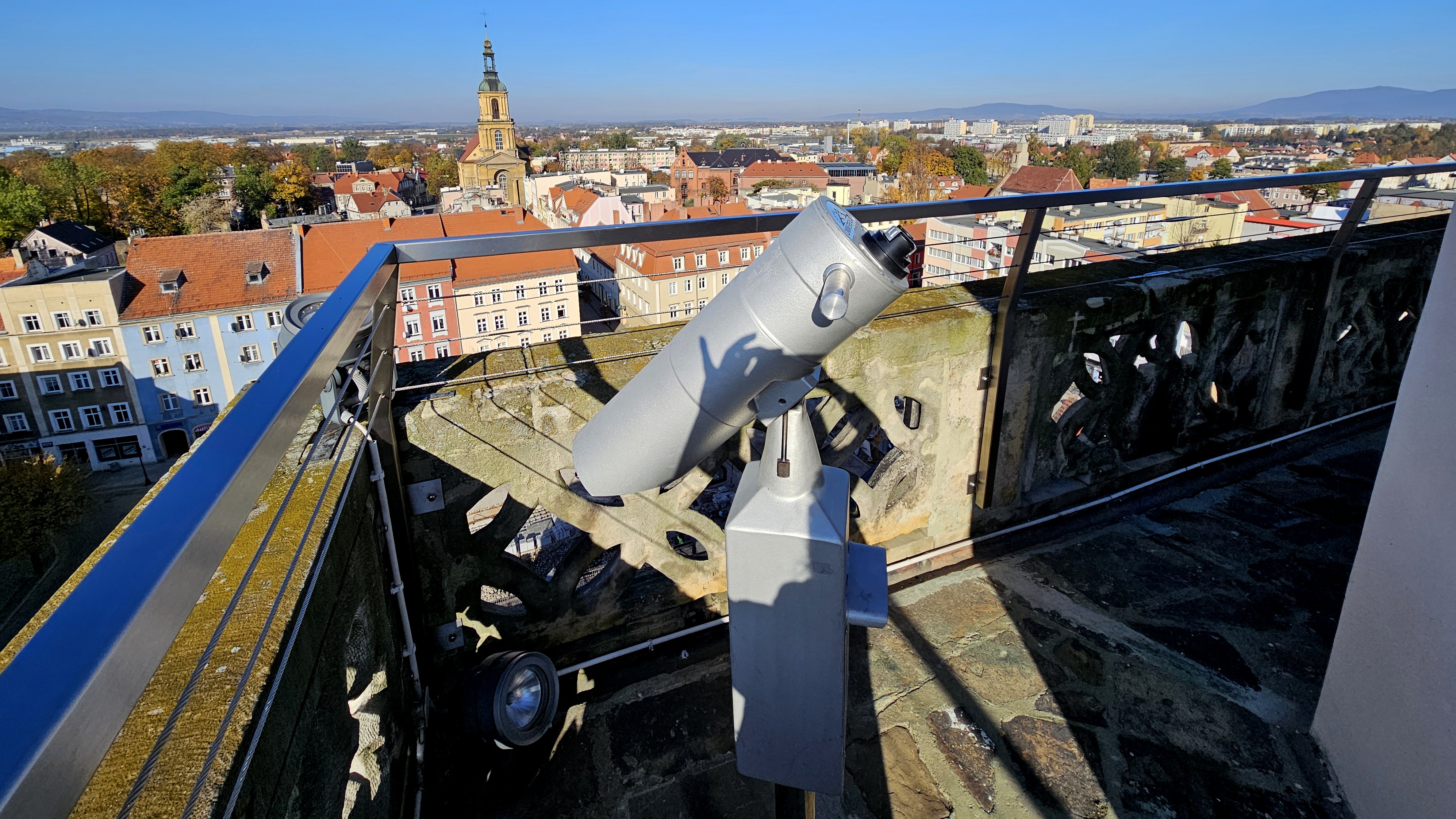 Wieża ratuszowa z kamiennymi i metalowymi barierkami, na pierwszym planie luneta, w tle panorama miasta