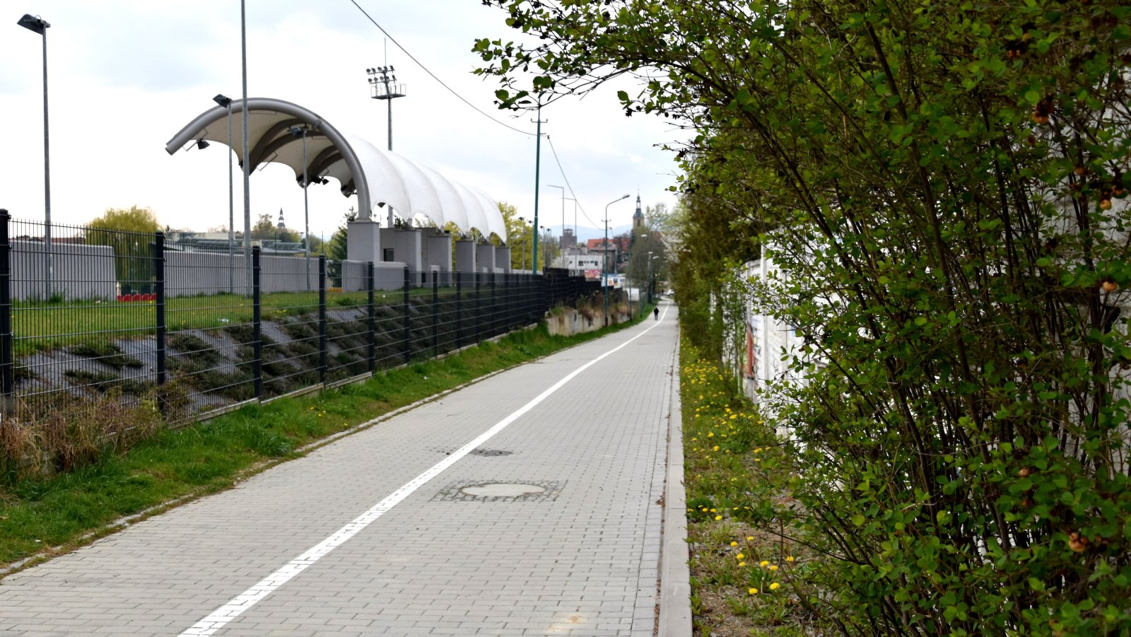 Chodnik i ścieżka rowerowa z kostki betonowej, po lewej za płotem zadaszenie trybun stadionu, po prawej zieleń