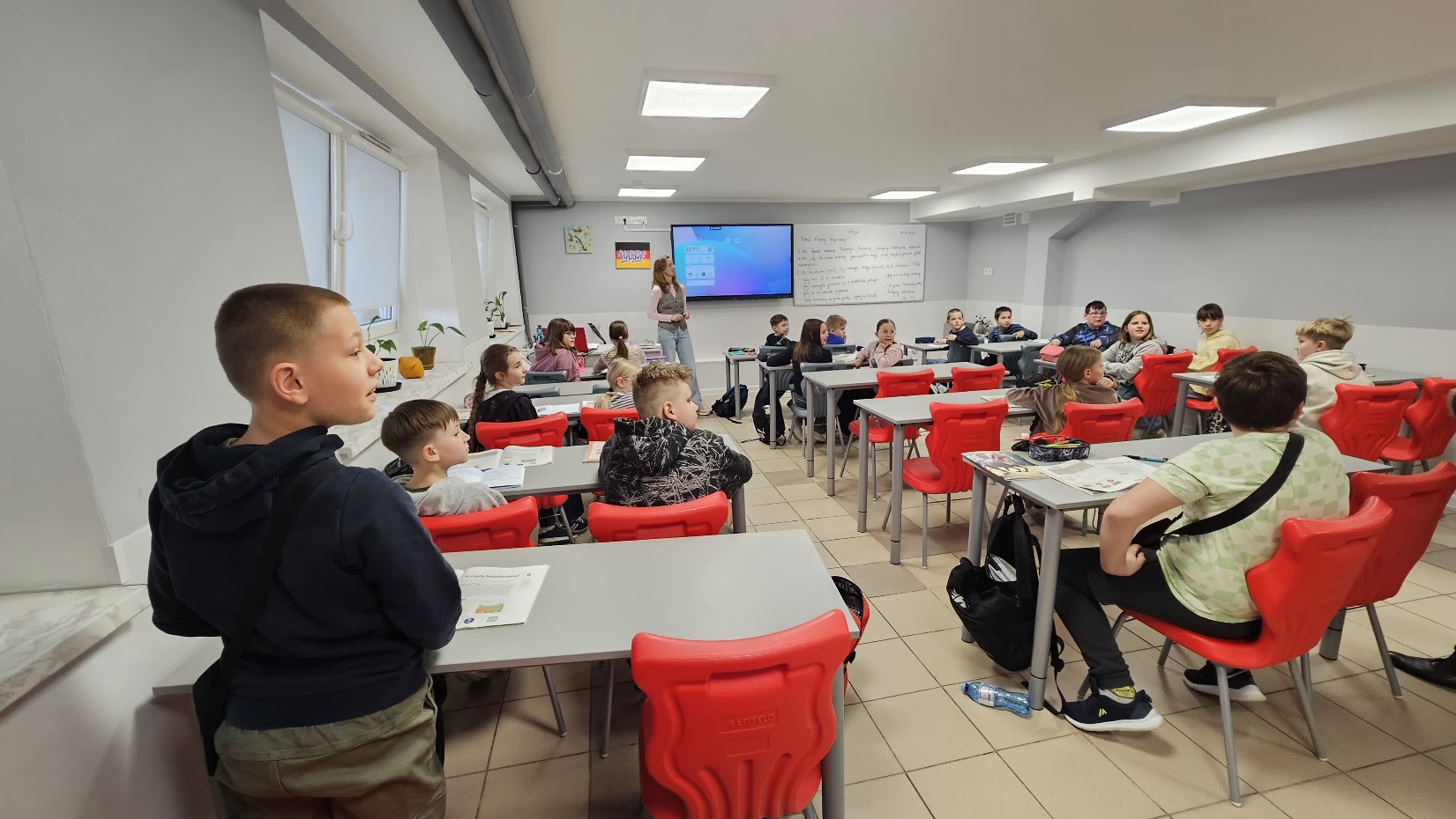 Klasa szkolna, białe ławki, czerwone krzesła, na ścianie tablica multimedialna