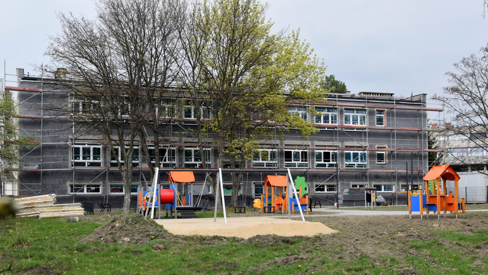 Plac zabaw z urządzeniami i piaskową nawierzchnią, w tle budynek w trakcie remontu