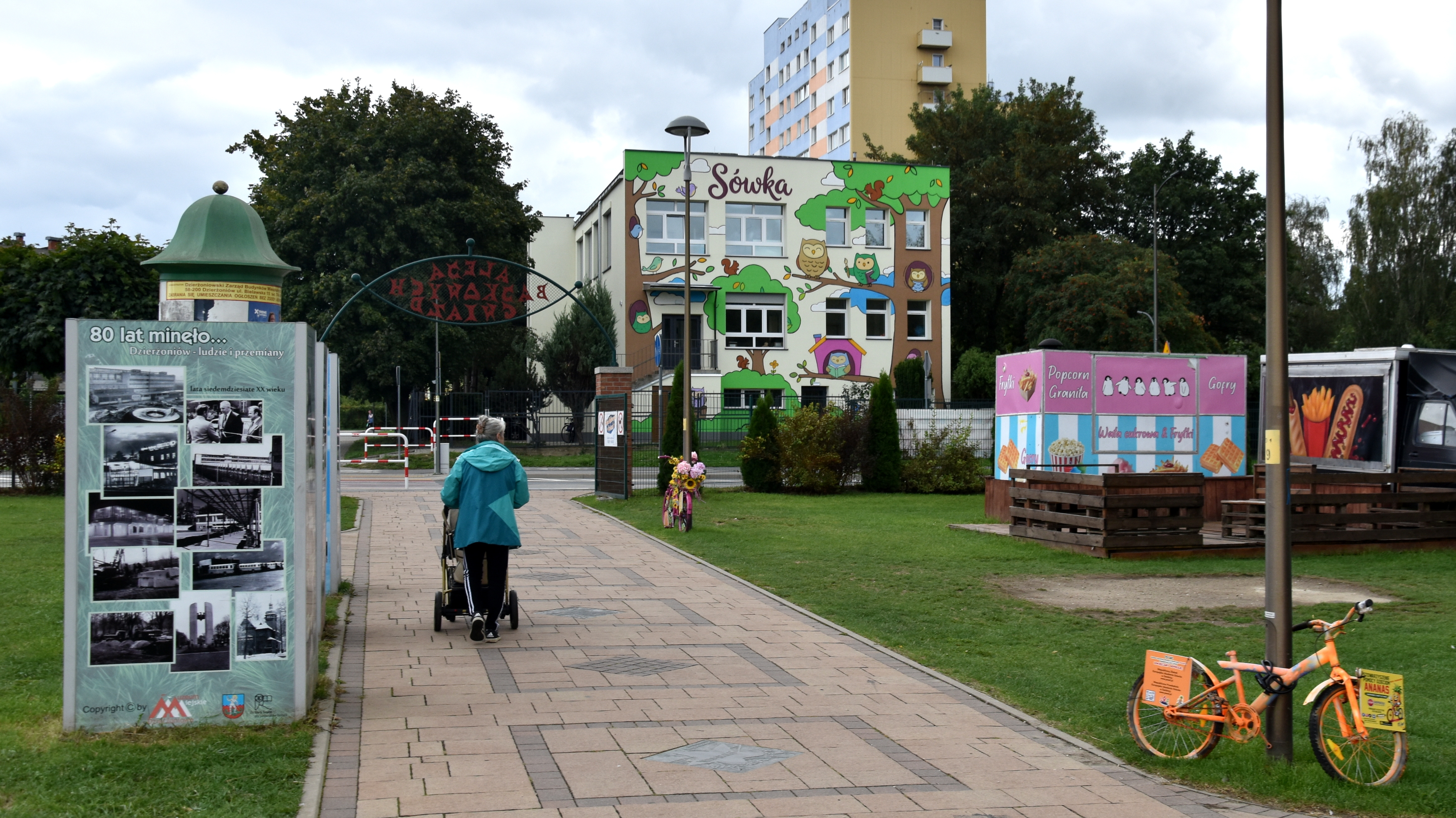 Kobieta prwadząca wózek, idąca aleją prowadzącą do żłobka, na ścianie budynku kolrowy mural z sowami
