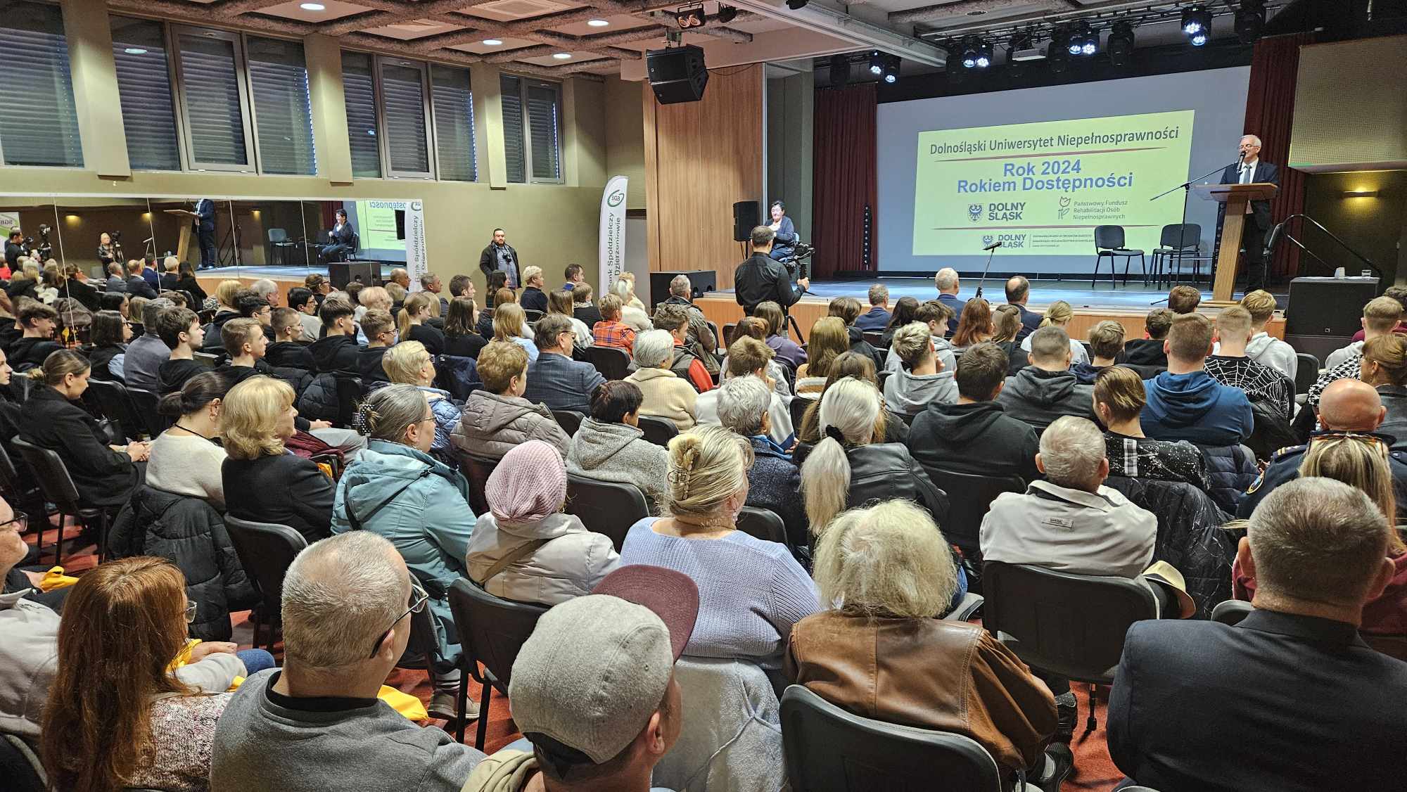 Duża sala wypełniona ludźmi siedzącymi na krzesłach, przy mównicy na scenie stoi mężczyzna