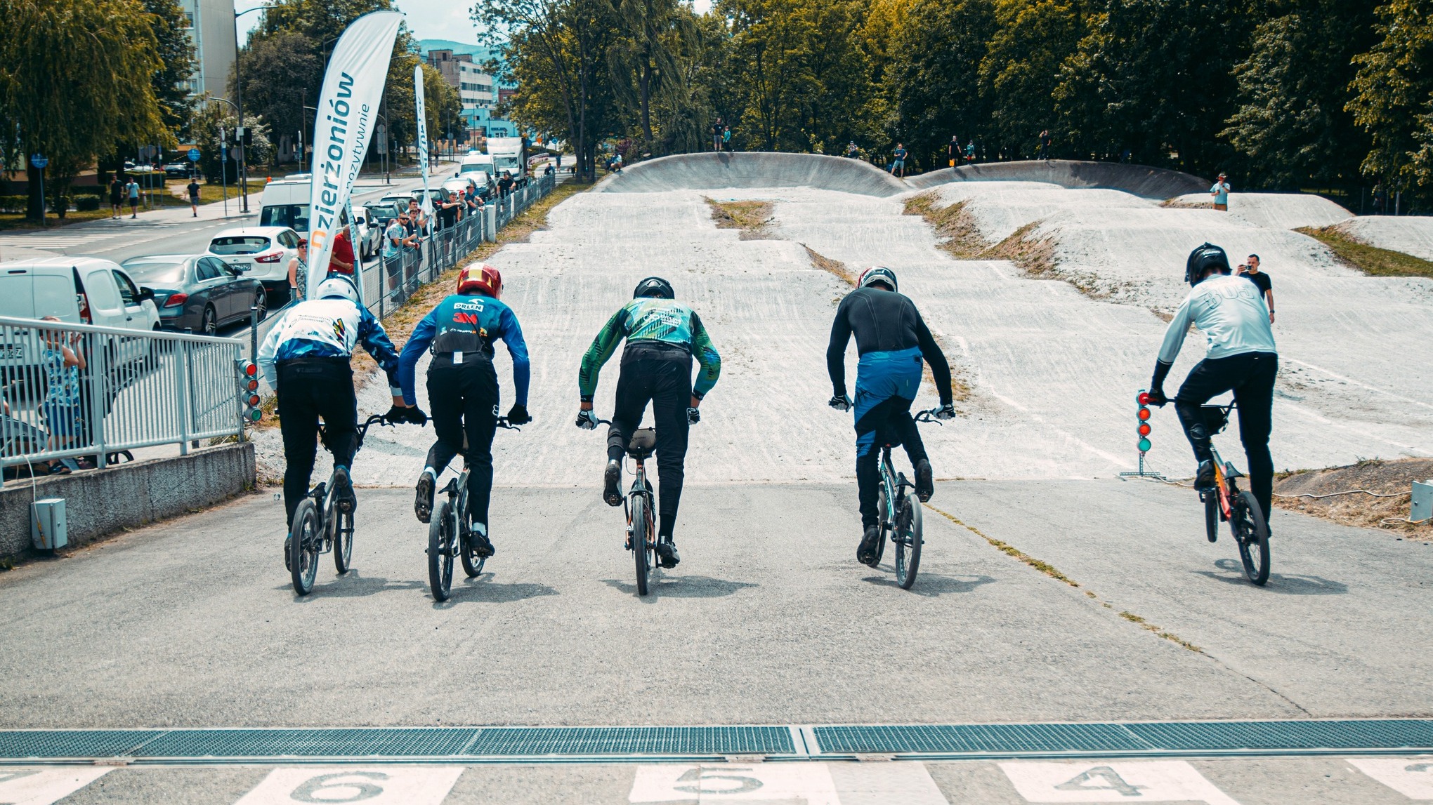 Pięciu rowerzystów jadących po pochylni , przed nimi tor wyścigowy, obok ulica, na chodniku kibice