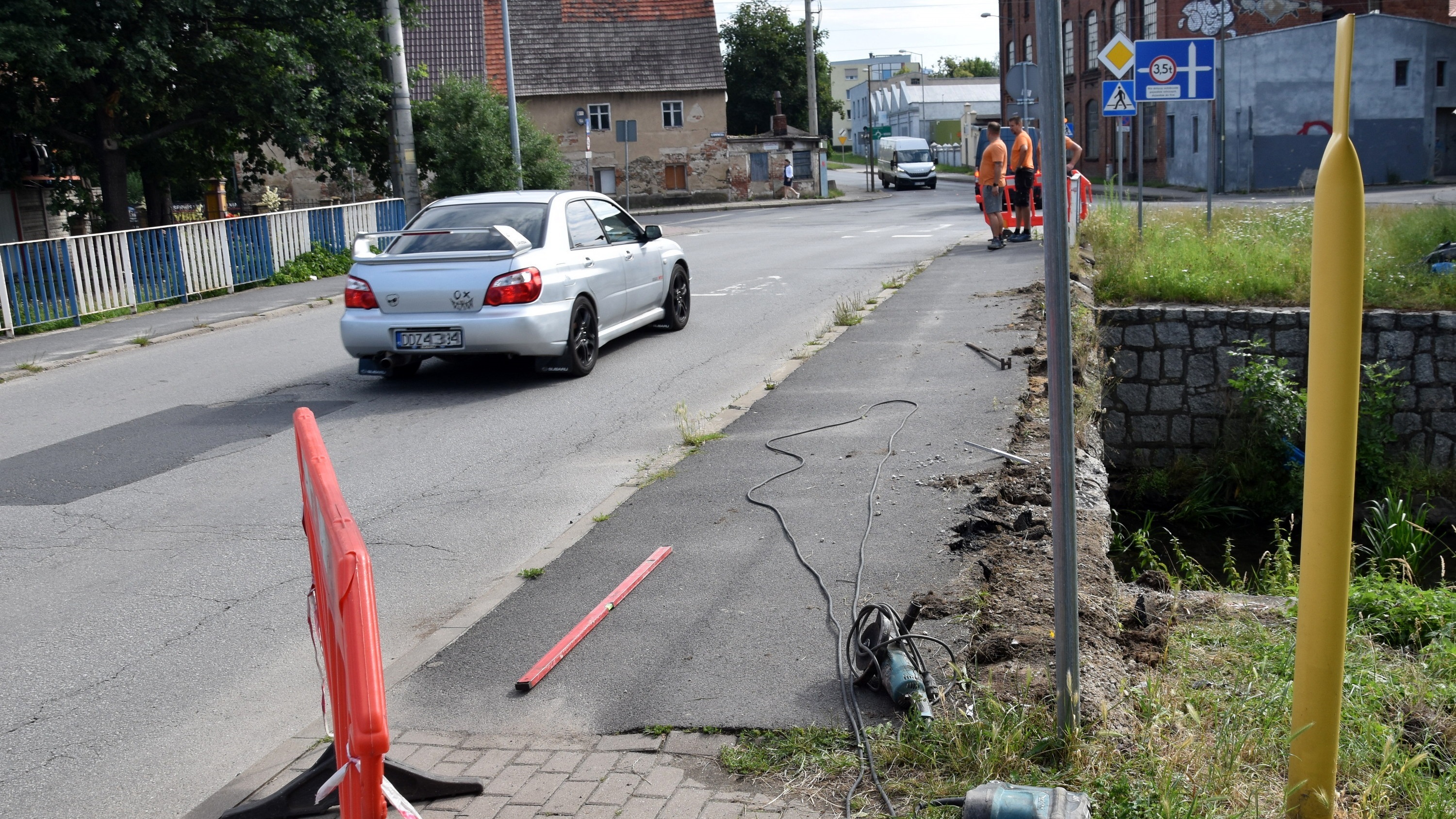 Samochód jadący po moście, na chodniku leża narzędzia, obo trzech pracowników remontujacych balustradę