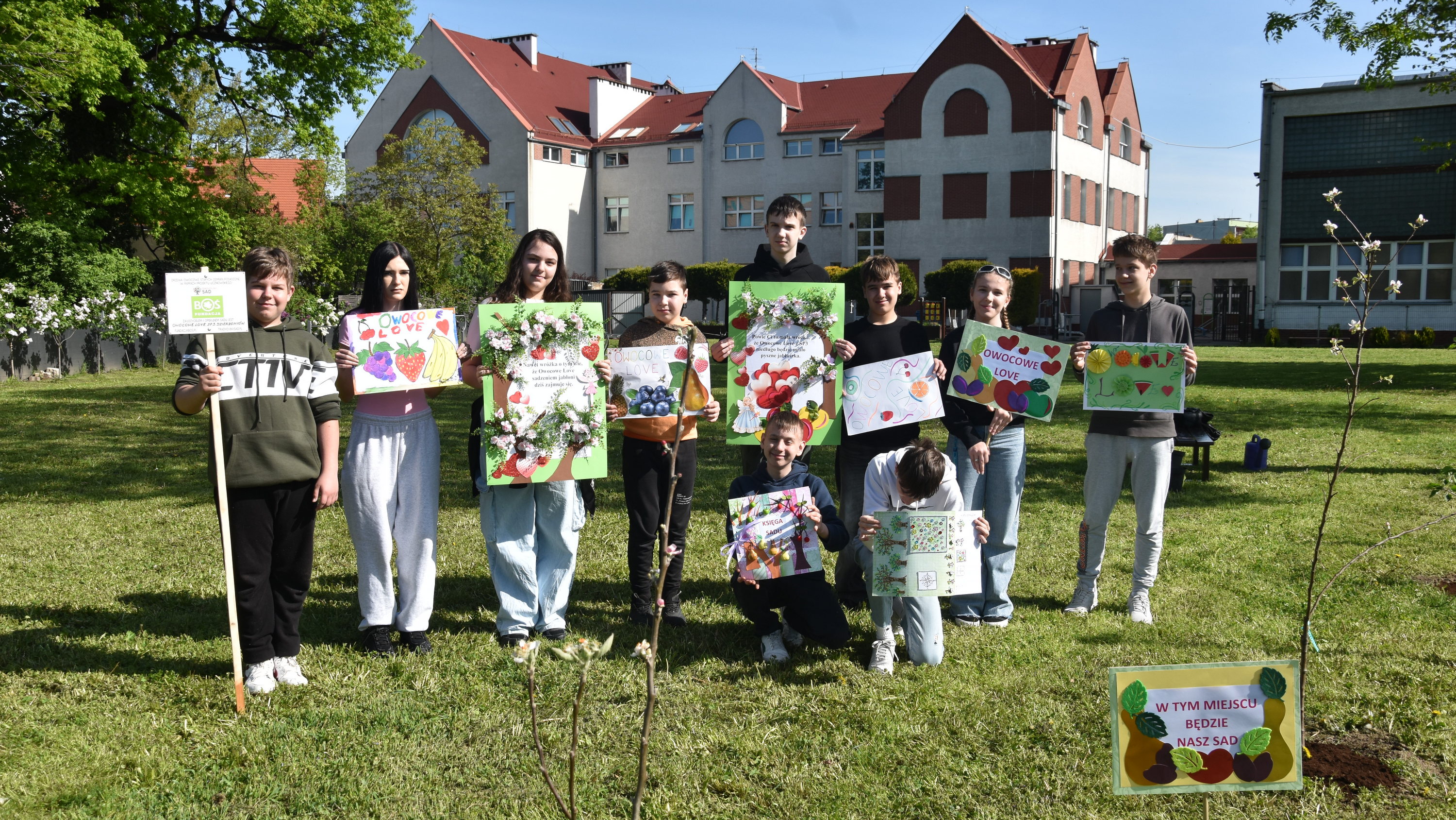 Dziesięcioro uczniów z kolorowymi planszami w rękach na tle budynku szkoły