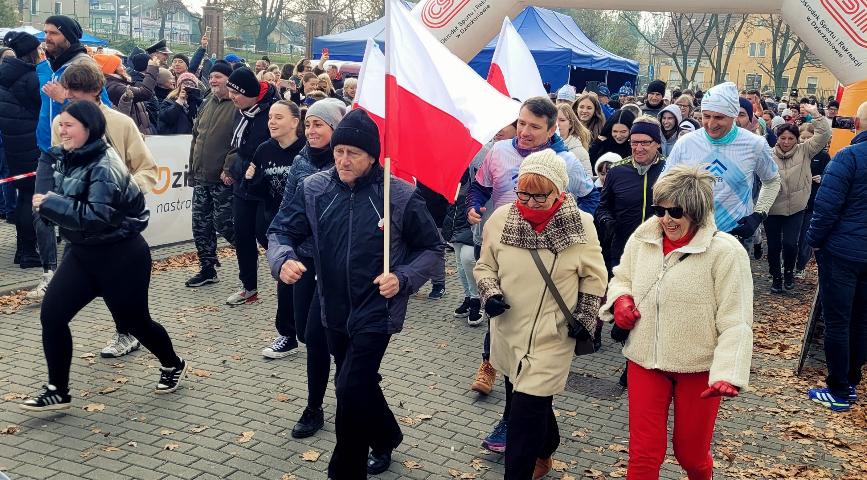 Grupa mieszkańców, niektórzy z flagami, startująca w biegu