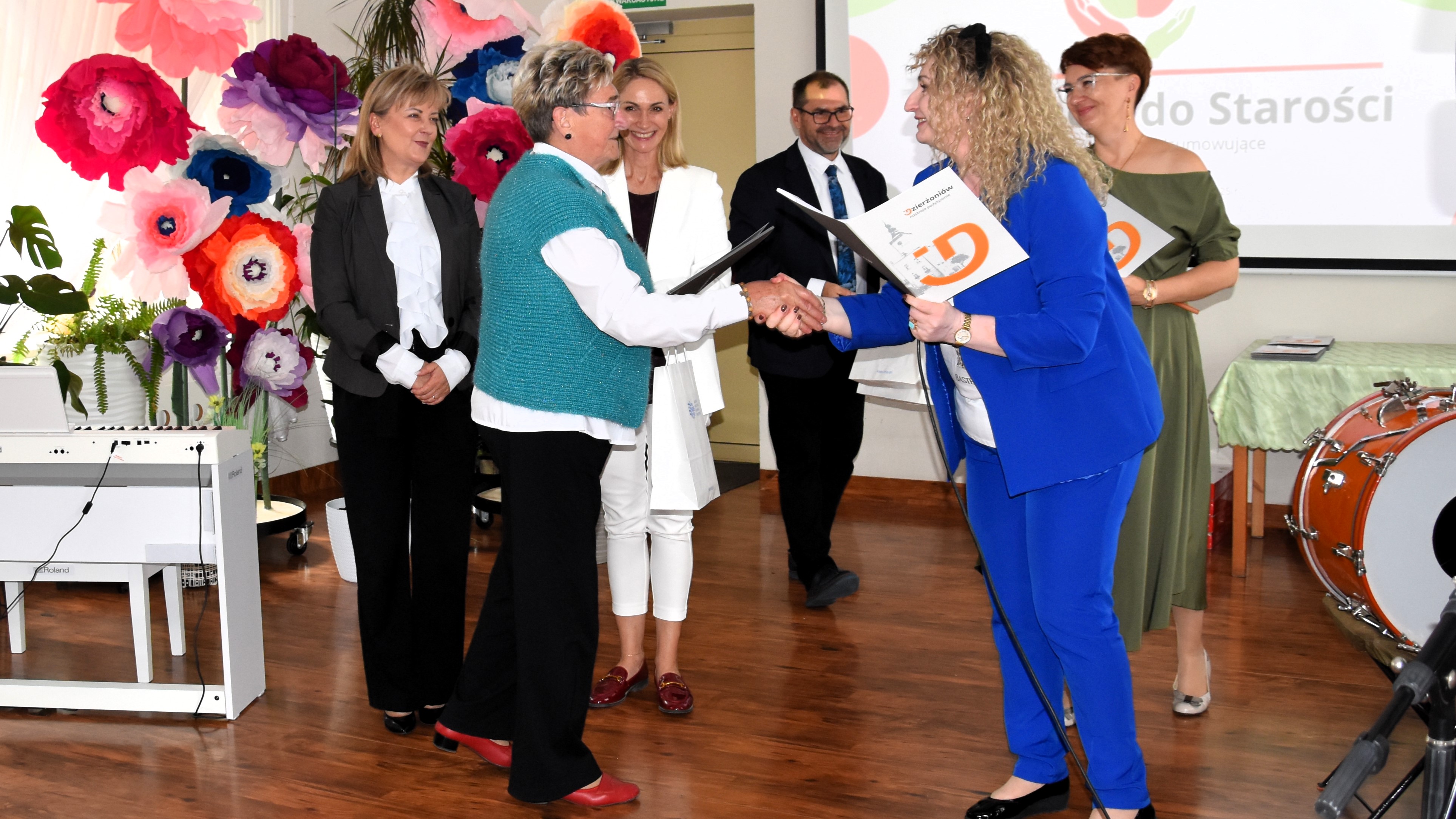 Kobieta wręczająca list gratulacyjny seniorce, na drugim planie trzy kobiety i mężczyzna