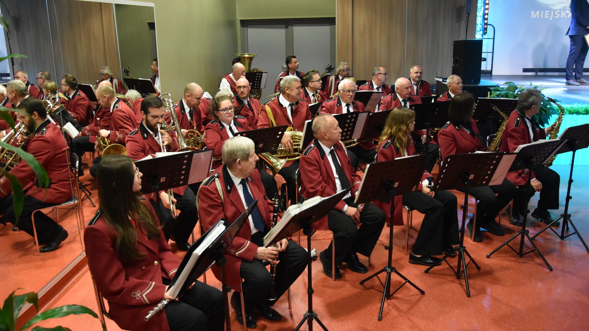 Muzycy orkiestry dętej siędzący na krzesłach, przed nimi nuty