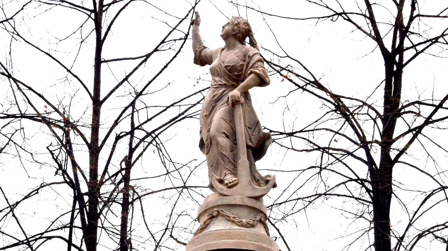 Rzeźba przedstawiająca kobiete w sukni z uniesioną głową i uniesioną jedną ręką