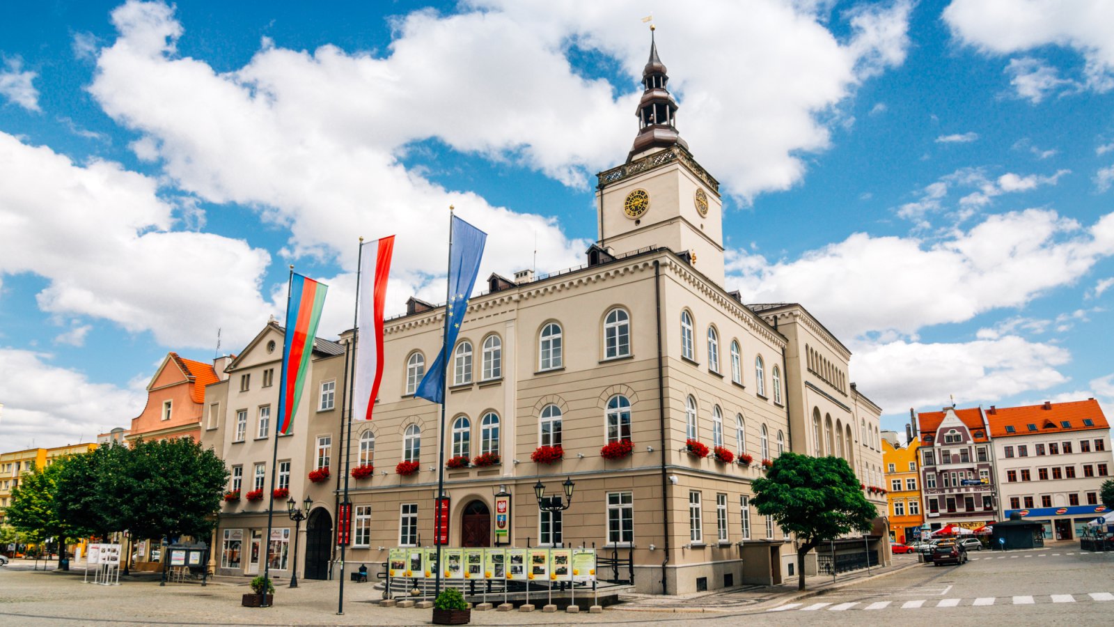 Budynek tarusza, przed nim flagi miasta, Polski i Unii Europejskiej, słoneczny dzień 