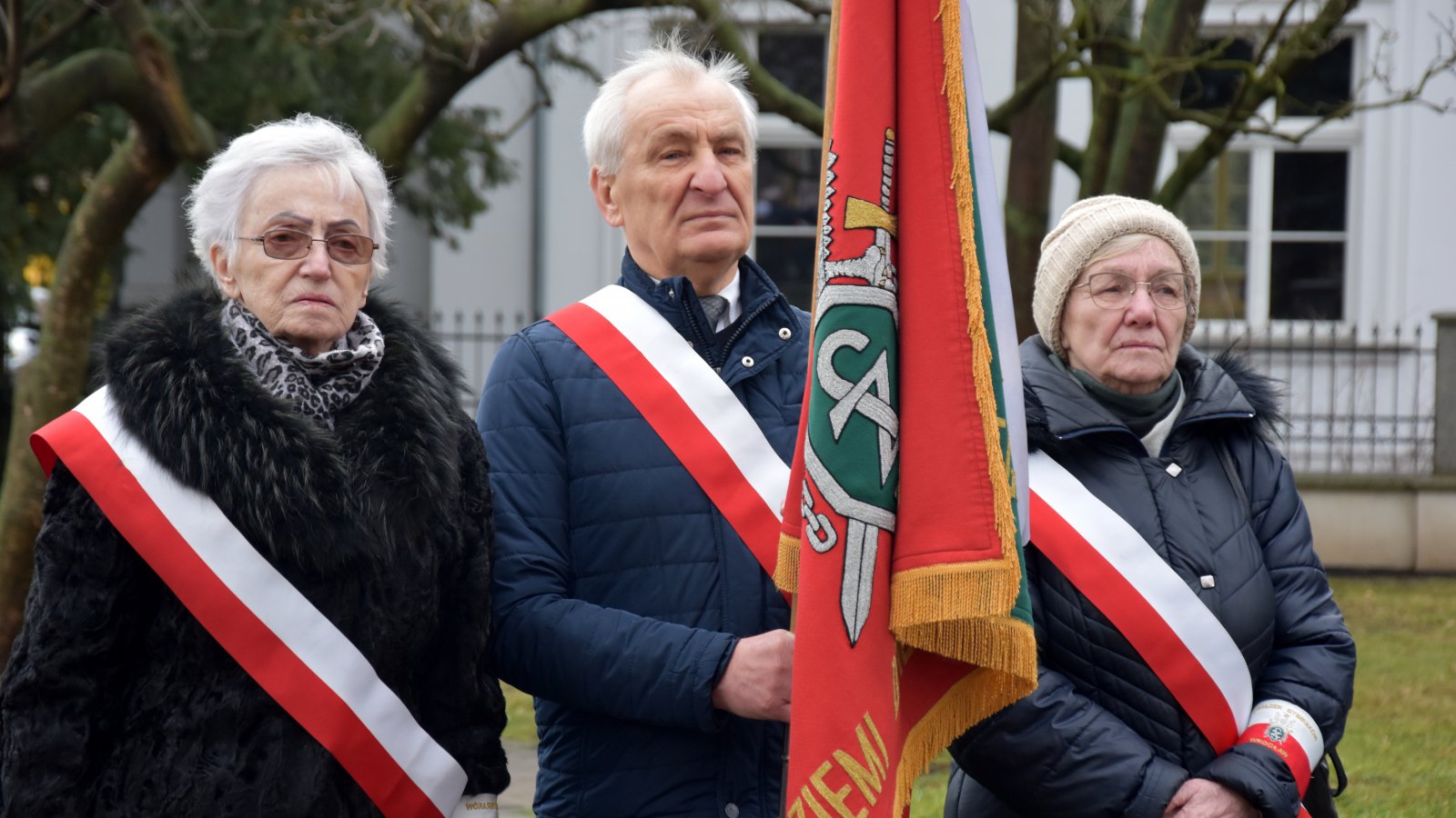Poczet sztandarowy, dwie starszze kobiety i jeden mężczyna trzymający flagę