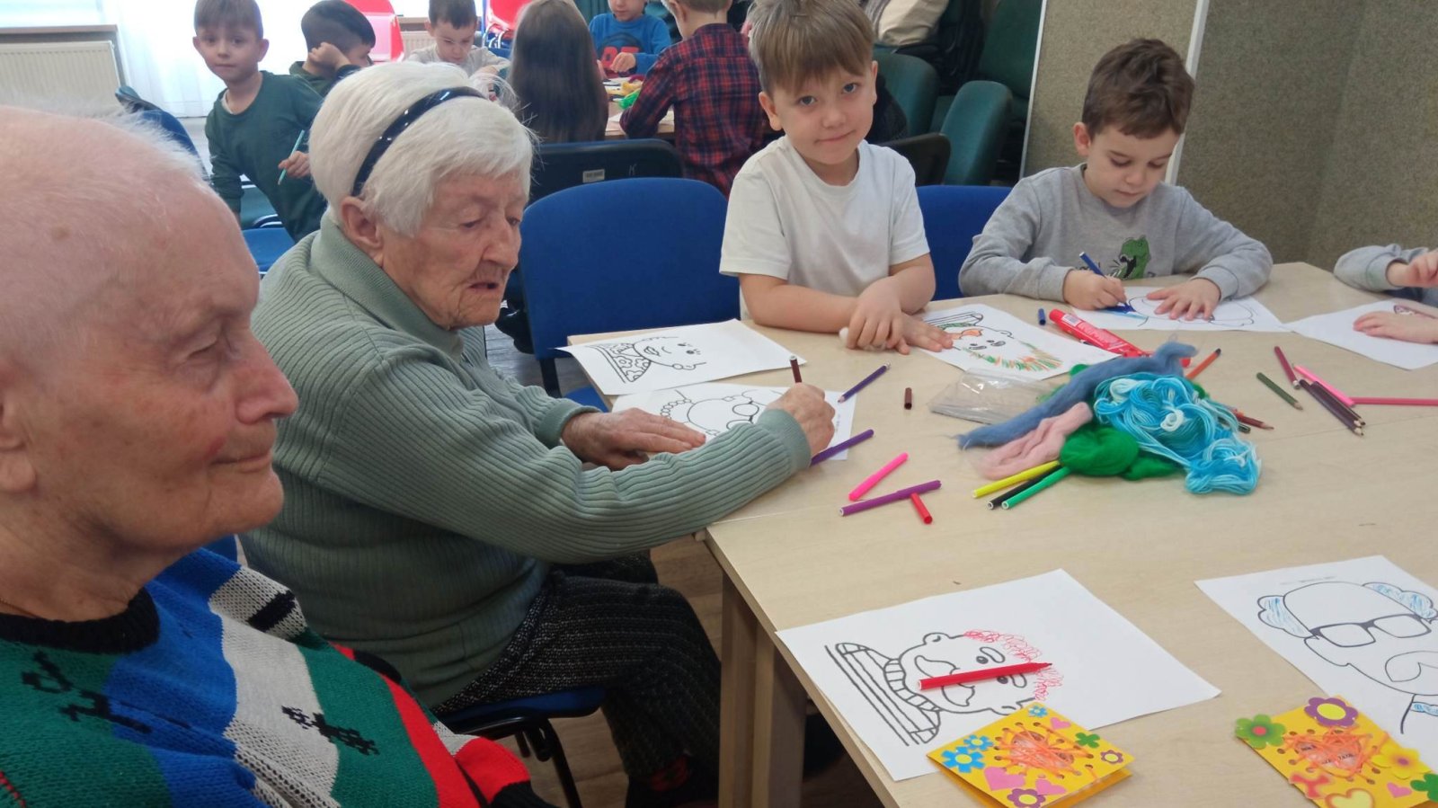 Seniorzy i dzieci przy jednym stole z kolorowankami.