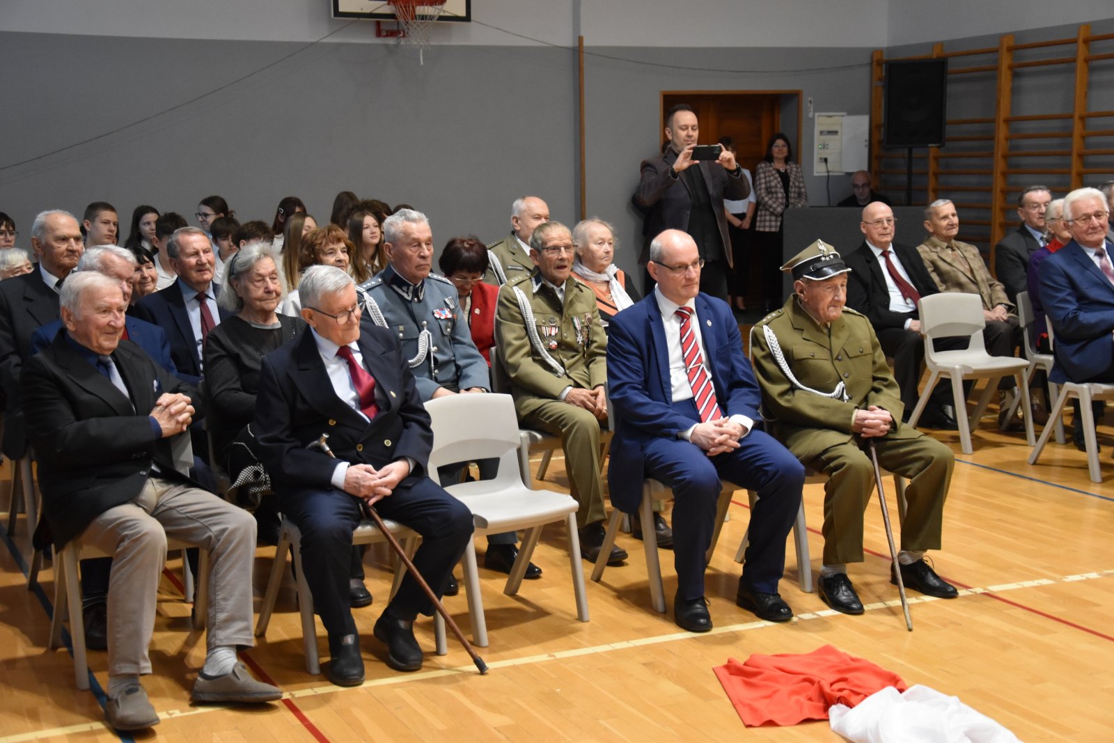 Kombatanci, niektórzy w mundurach siedzący na krzesłach, z tyłu grupa młodzieży