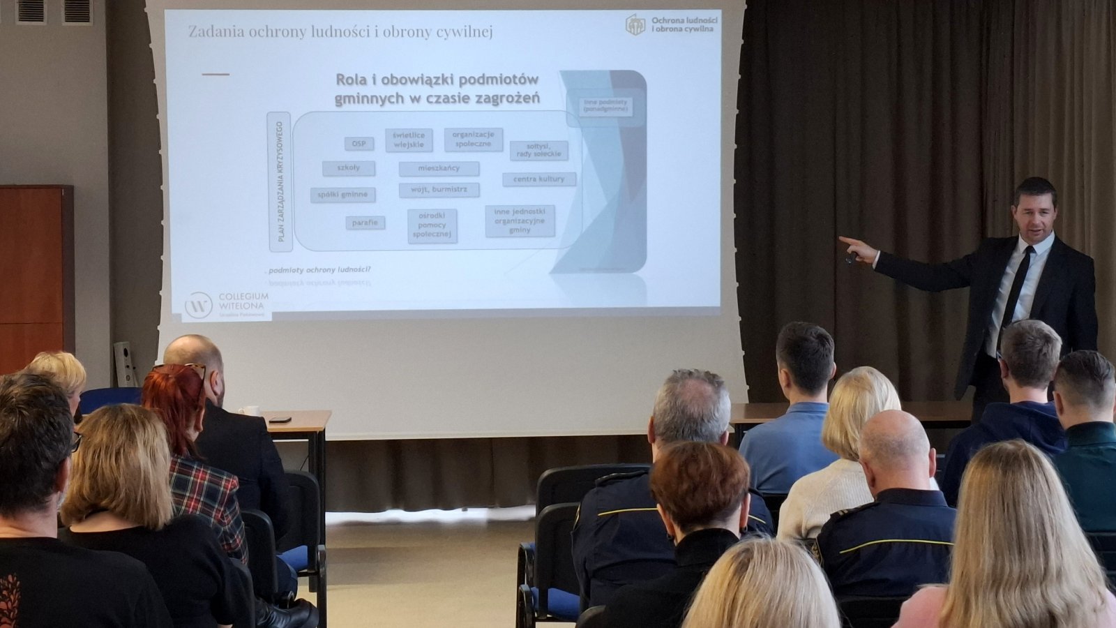 Szkolenie mężczyzn w garniturze wskazuje na ekran, na pierwszym planie kilkunastu uczestników siedzących na krzesłach