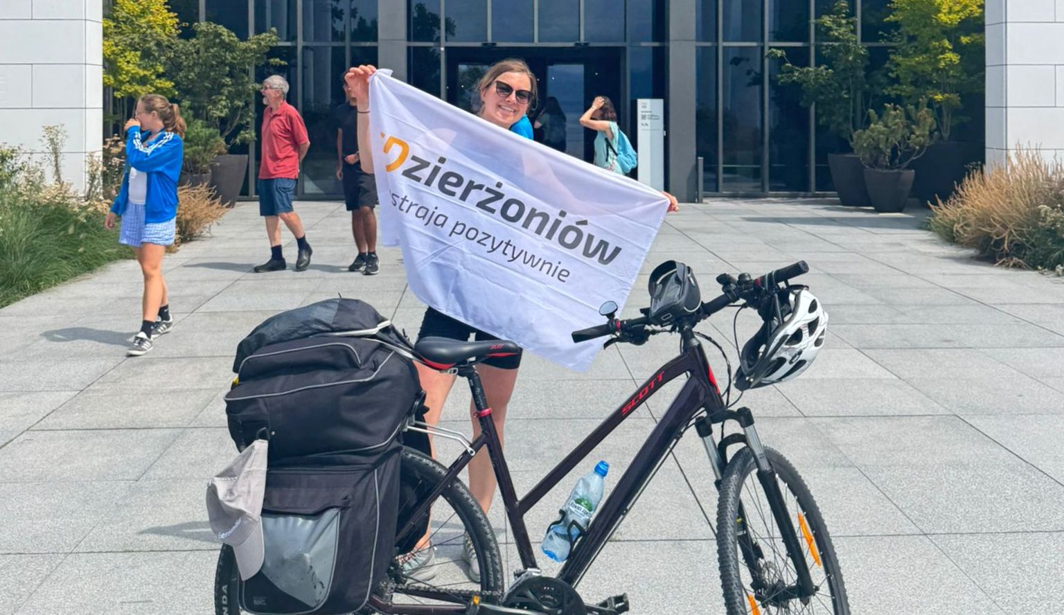 Uśmiechnięta dziewczyna trzyma flage z napisem Dzierżoniów, przed nią rower