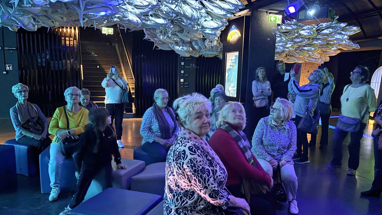 Seniorki siedzące i stojące w dużym pomieszczeniu, niebieskie światło