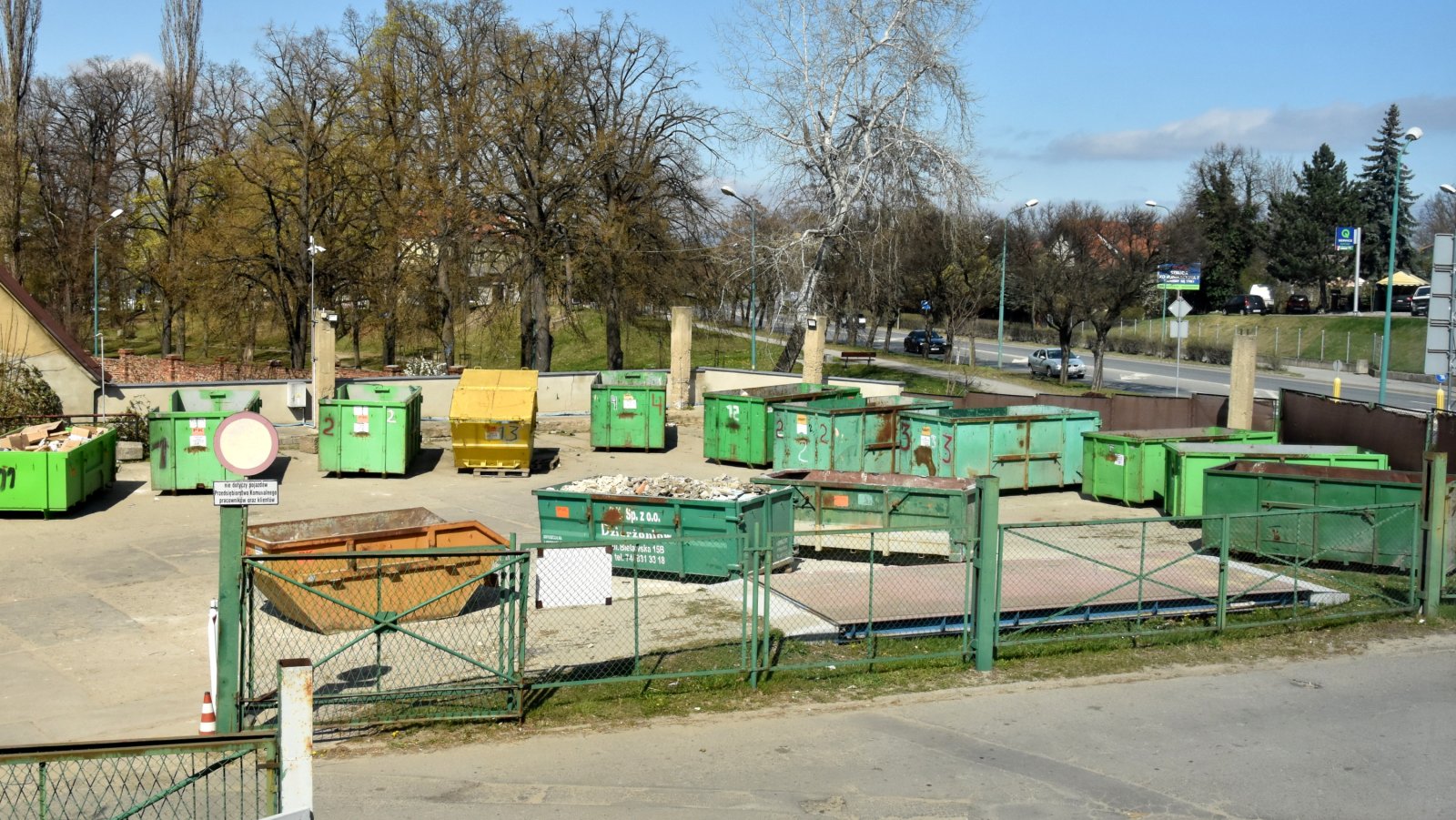 Duże zielone kontenery na śmieci ustawiione na placu, obok droga