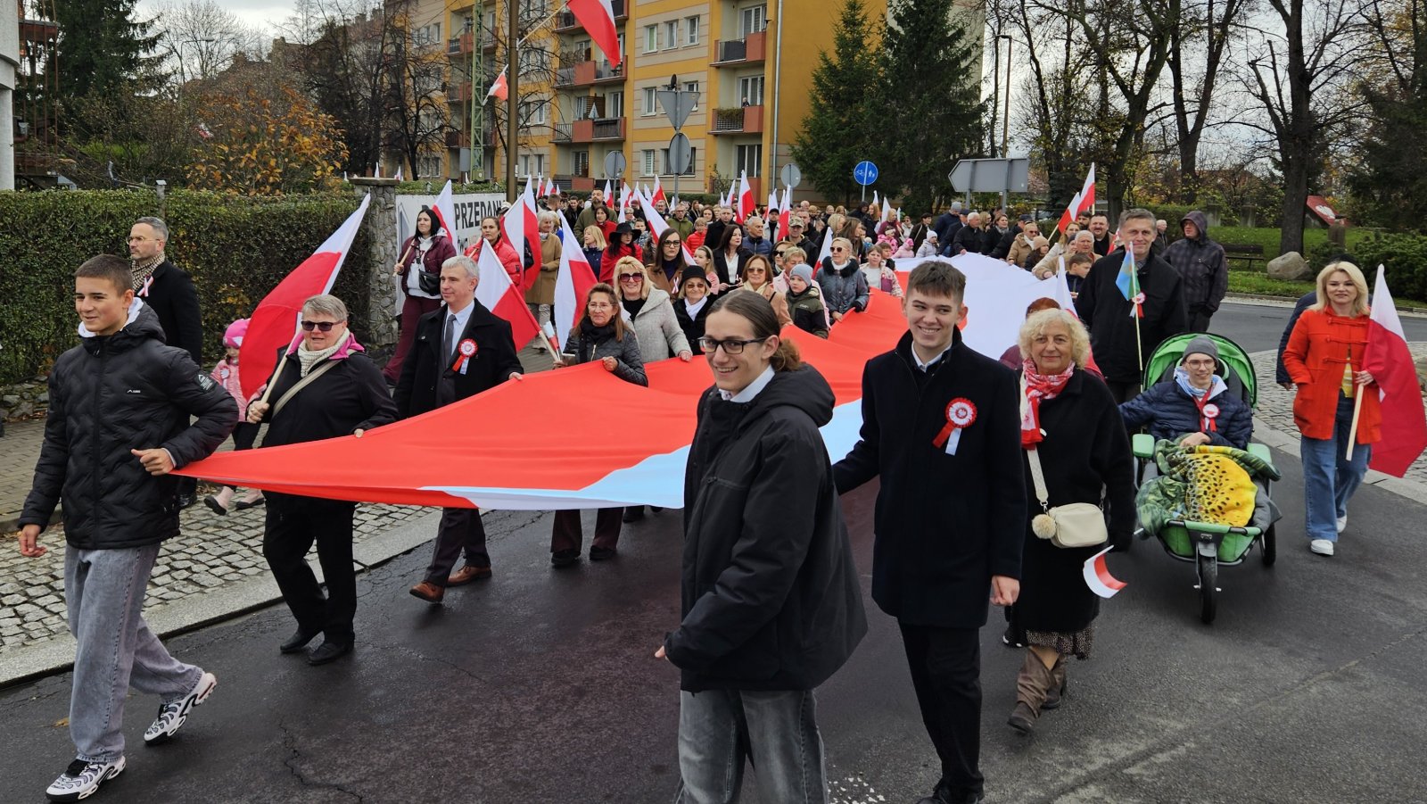 Tłum mieszkańców idących ulicą i niosących długą biało-czerwoną flagę