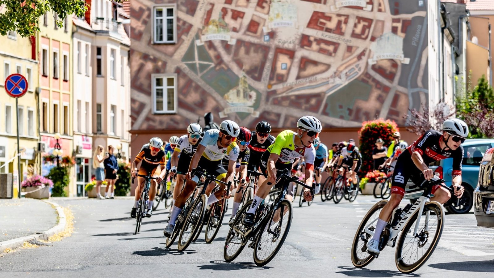 Kolarze jadący ulicą, w tle budynek z muralem