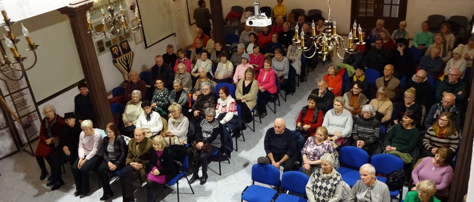 Wnętrze synagogi, kilkadziesiąt osób siedzi na krzesłach