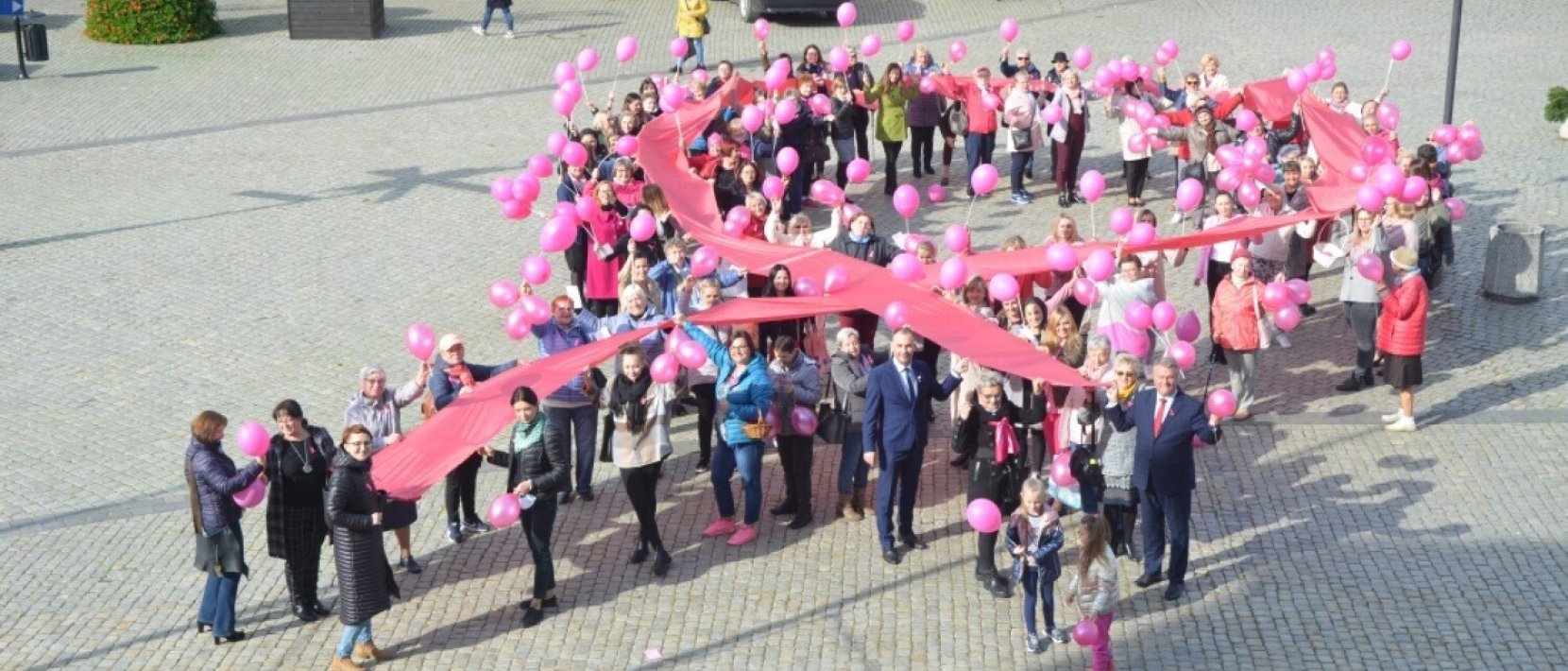 Zdjęcie z lotu ptaka. Duża grupa grupa ludzi trzymająca ogromną różową wstęgę oraz różowe balony w rynku dzierzoniowskim
